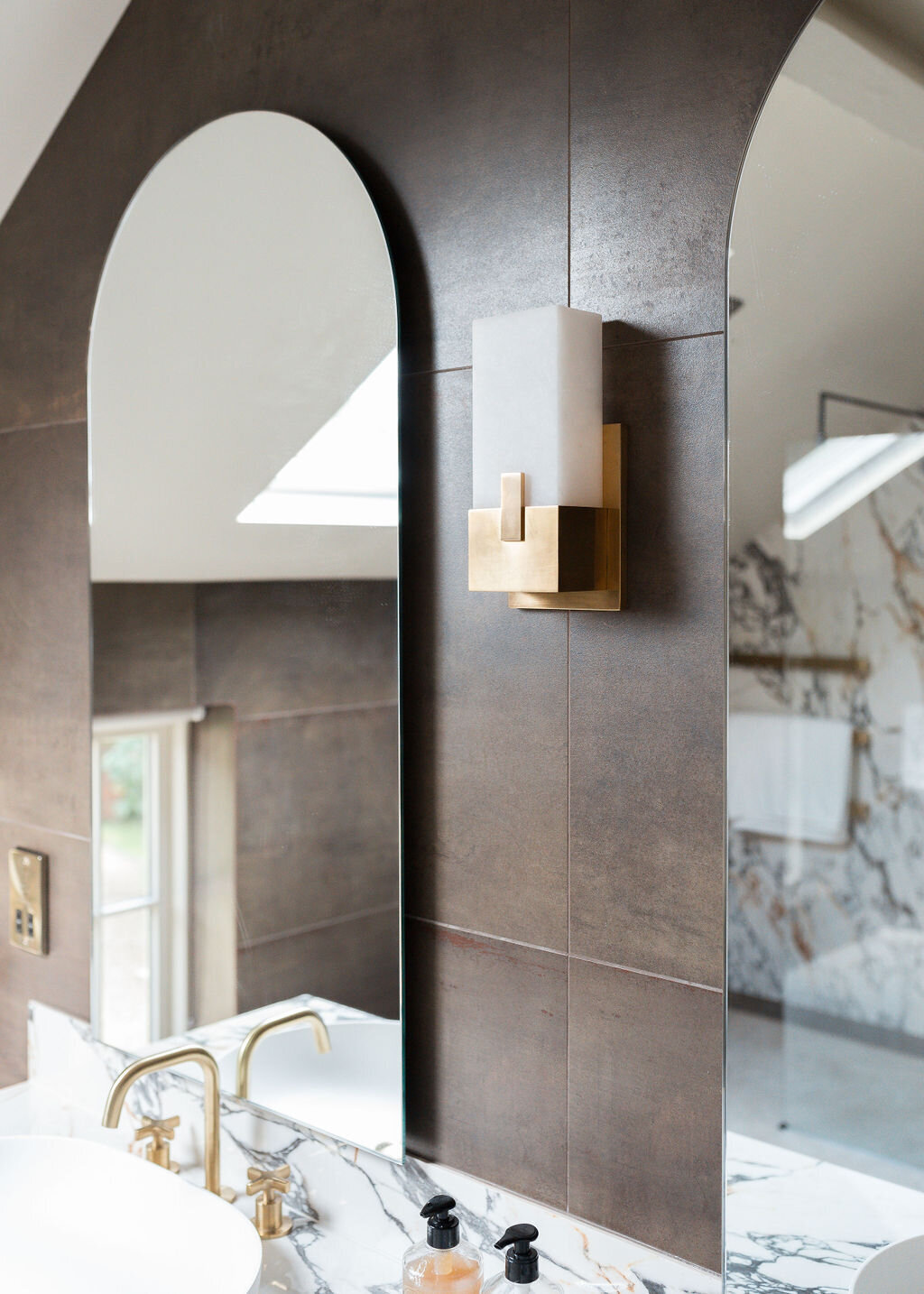 Two arched mirrors are mounted above sinks in a modern bathroom. The room has dark tiled walls and marble countertops.