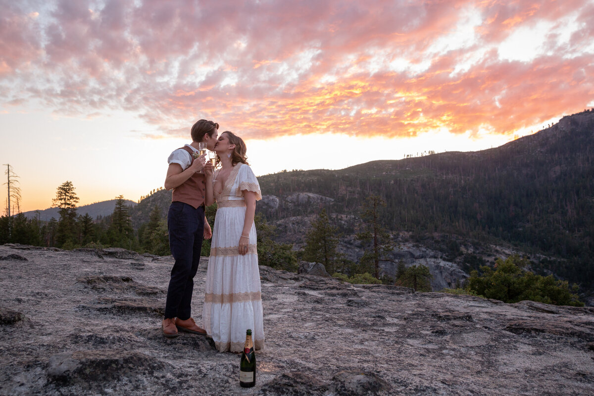 Two brides kiss while holding champagne glasses as the sky turns pink, orange, and purple behind them on their Yosemite elopement day.