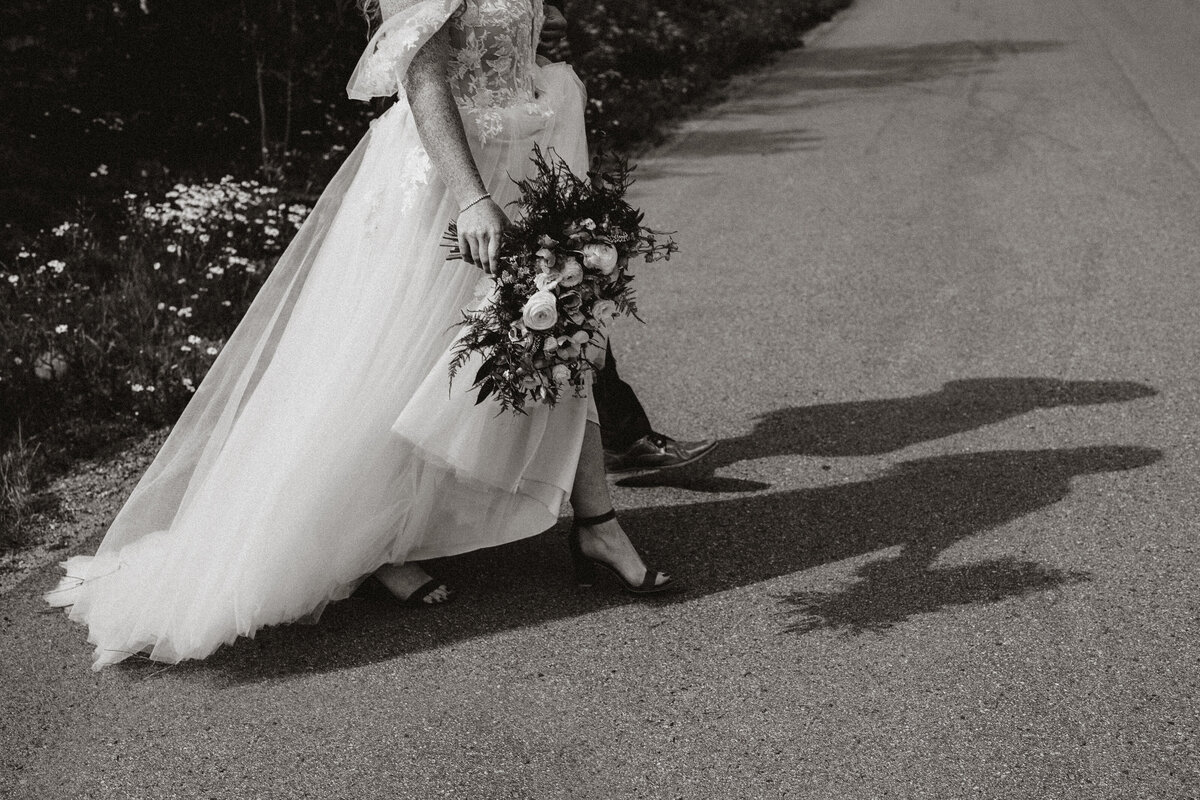Bride walks down mountain road holding bouquet, her shadow cast beside her during Breckenridge wedding.
