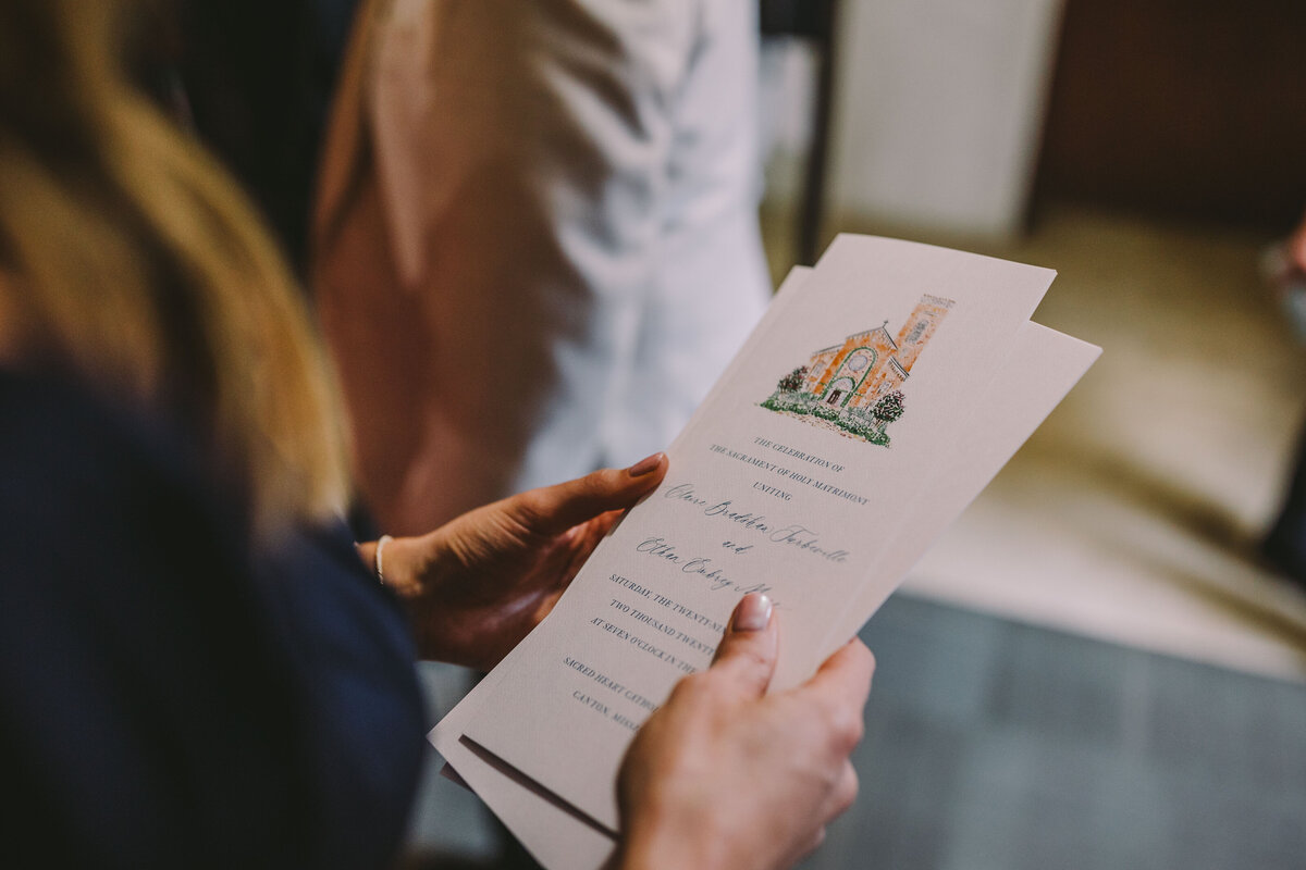 Guest holding wedding program featuring a watercolor illustration of the chapel at Sacred Heart in Canton, Mississippi.