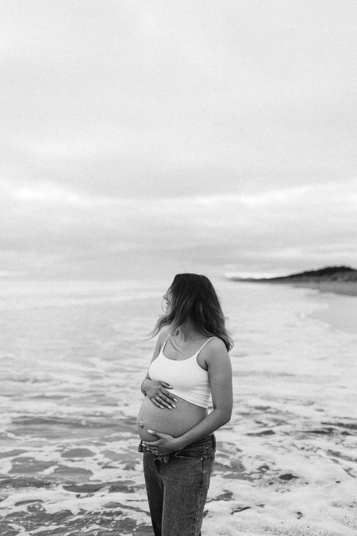 A mother to be wearing a white crop top and jeans standing in the waves during a maternity photoshoot at a popular christchurch beach with lucy jane photography