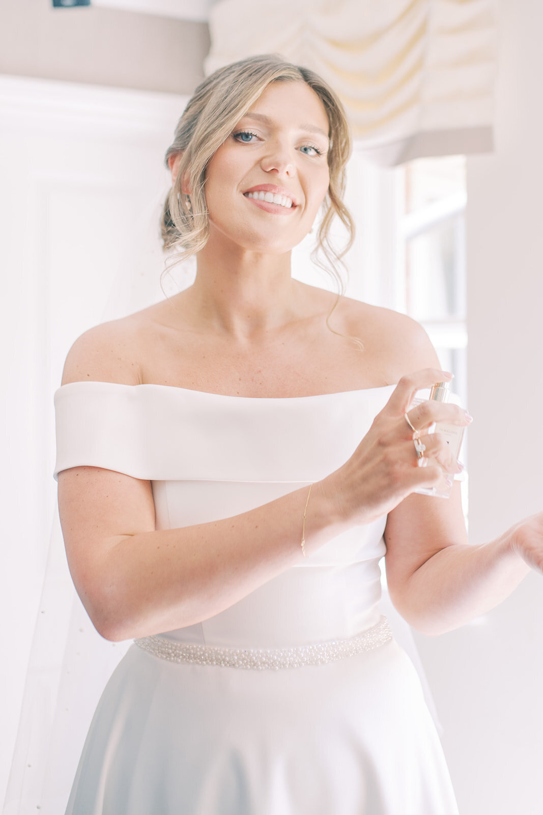 colour inmage, edited in a light and airy style. Bride is spraying her perfume on her wrist and is smiling at the camera