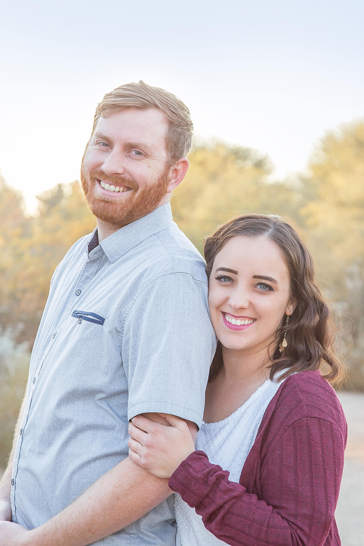 Happy redheaded man with brown haired wife snuggled standing behind him smiling