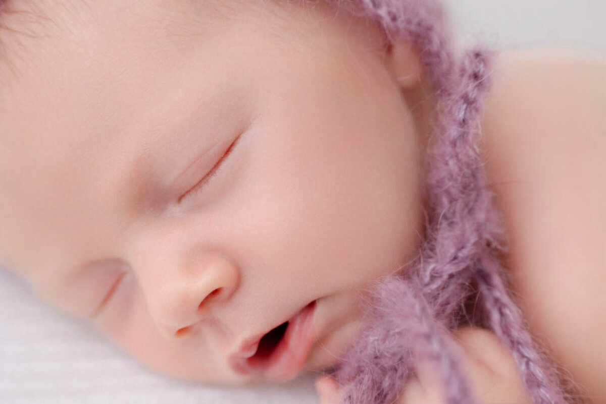 close up of two-week baby girl posed lying on white blanket and wearing a purple bonnet and covered in a lacy purple baby wrap.