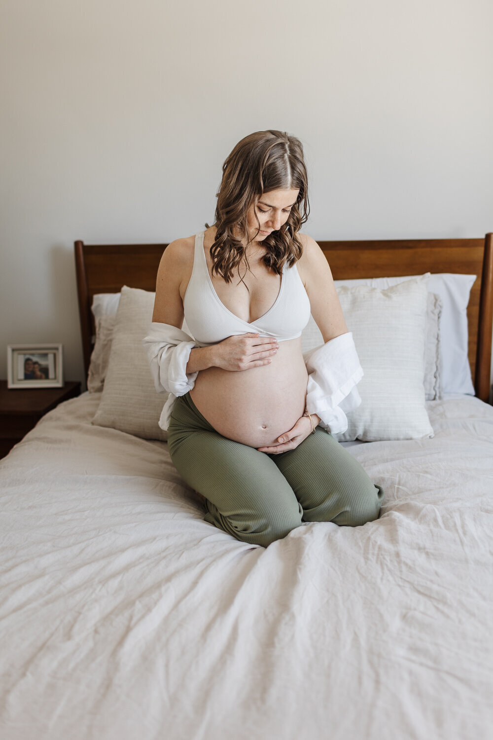 Pregnant woman dressed in white bralette and green lounge pants sitting on her bed admiring her belly