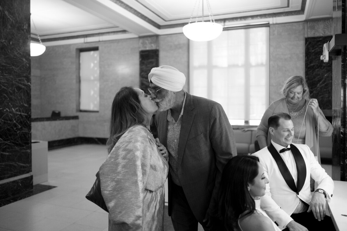 Black and white candid photo of family members sharing a kiss while waiting inside New York City Hall during Japna and Chris’s elopement, captured by NYC wedding photographer Perry Hancock.