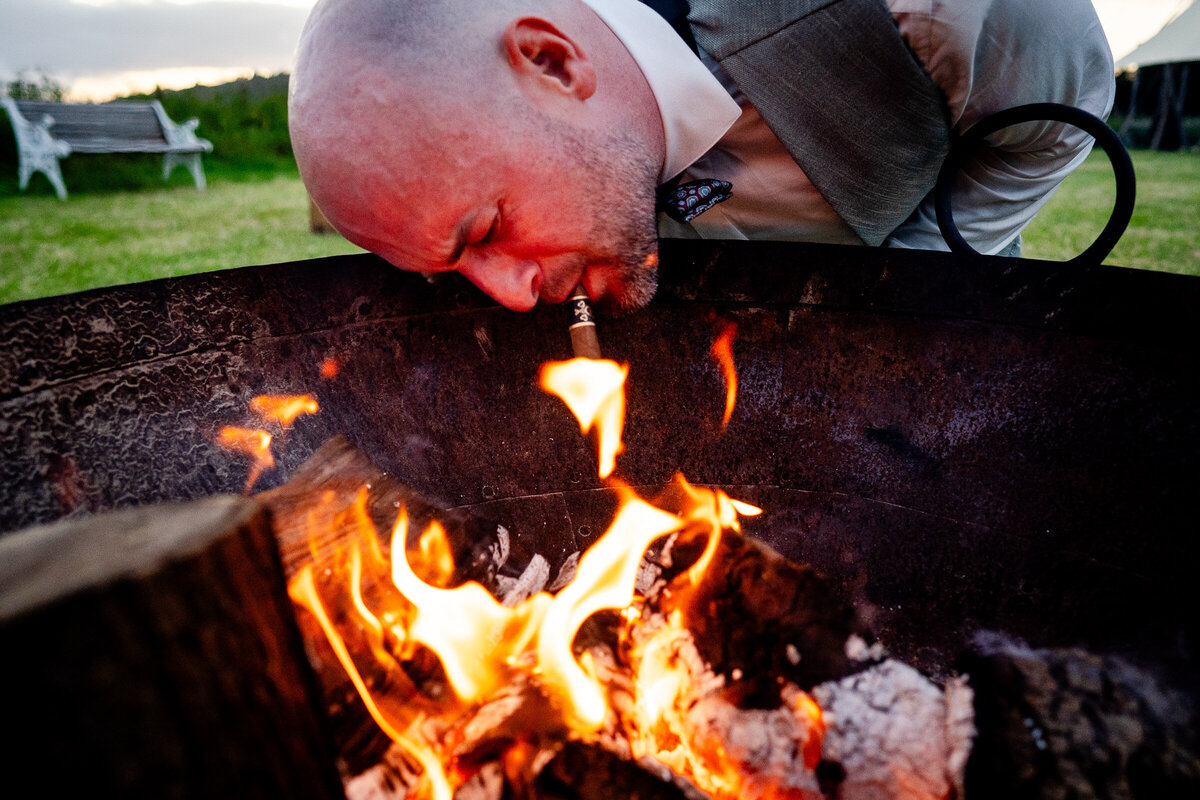 The groom is lighting his cigar in a fire pit with his head dangerously close to being singed