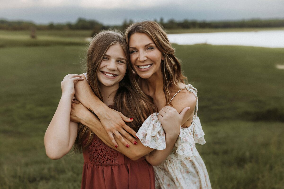 Two people smiling and embracing outdoors with a grassy field and a body of water in the background.
