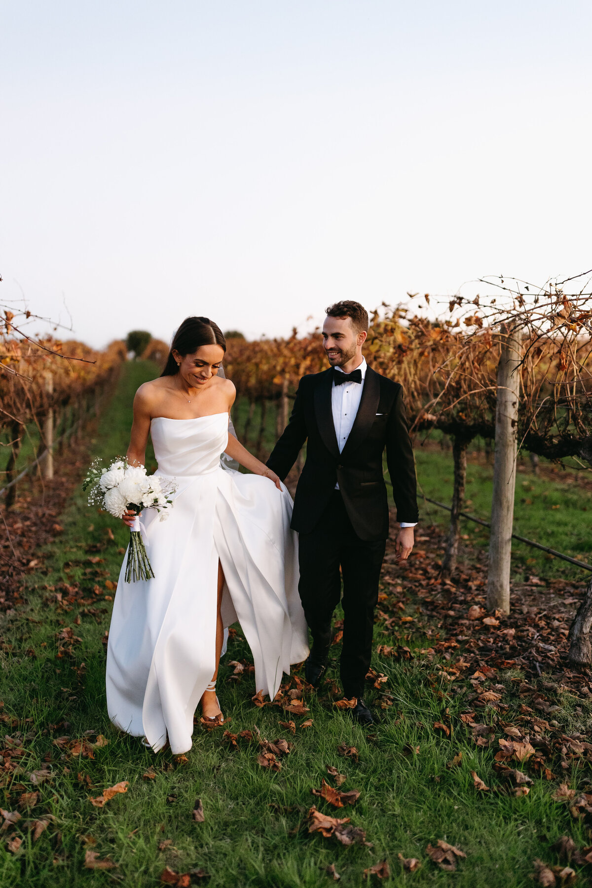 An image of a bride and groom in a leafy vineyard. The bride is holding her dress up. The groom is looking toward her and is supporting her as she walks through the vineyard.