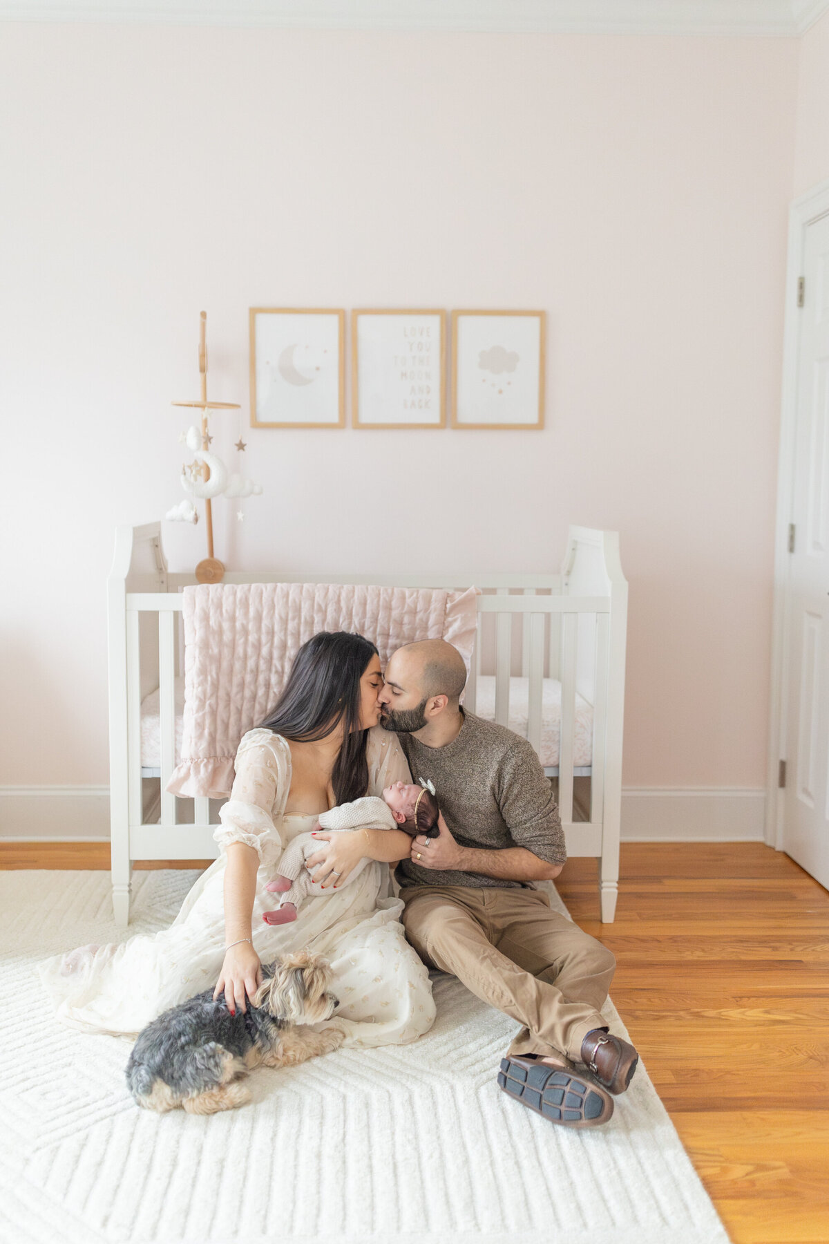 Husband and wife sitting together in front of their baby's crib, gently holding their newborn infant. The couple shares a tender moment, looking lovingly at their baby in the peaceful nursery setting