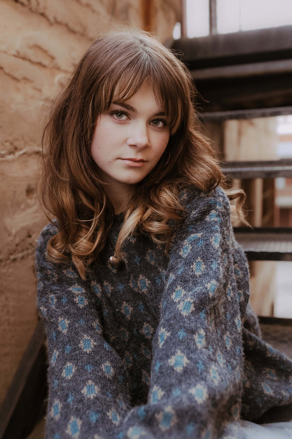 A woman with long brown hair sits on stairs, wearing a gray patterned sweater. She looks toward the camera with a neutral expression.