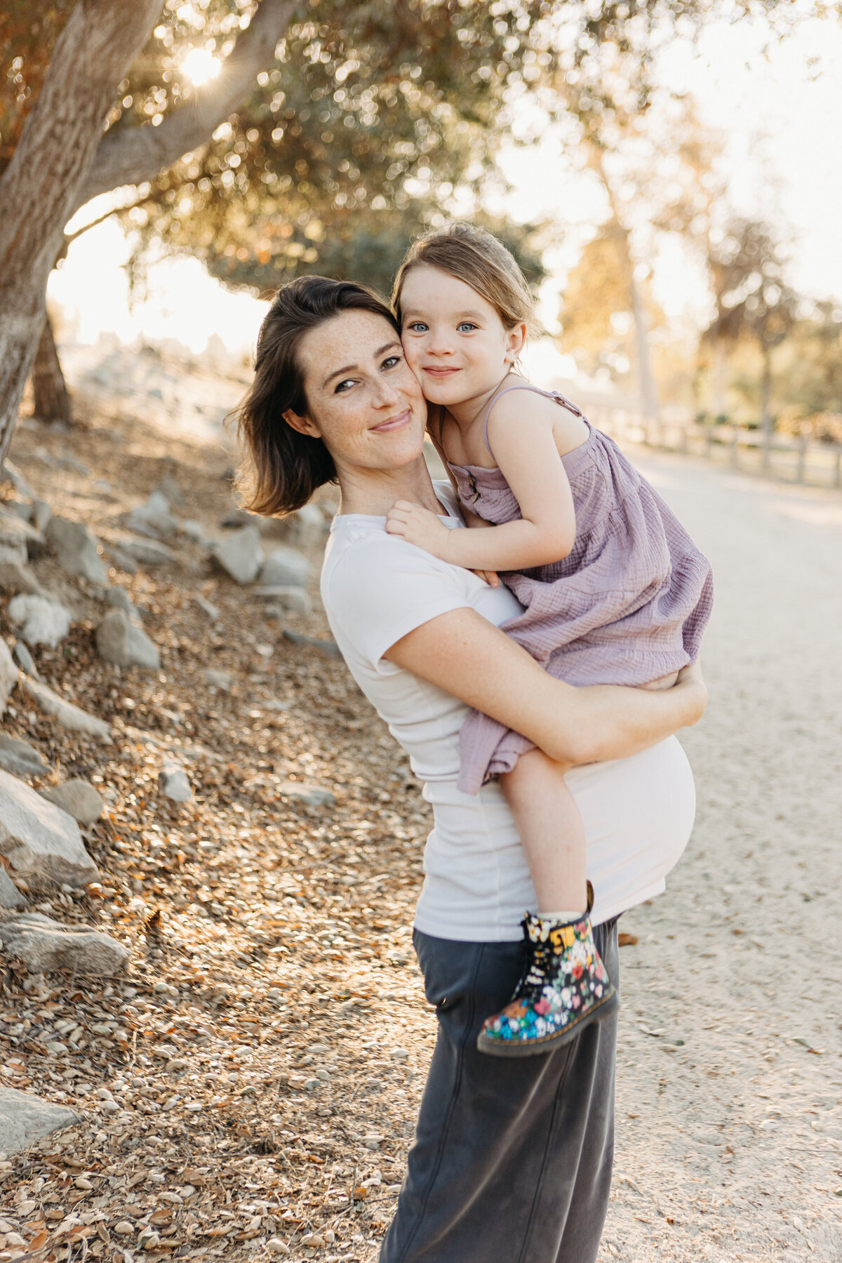 Daughter hugs pregnant mama with their cheeks pressed together at golden hour.