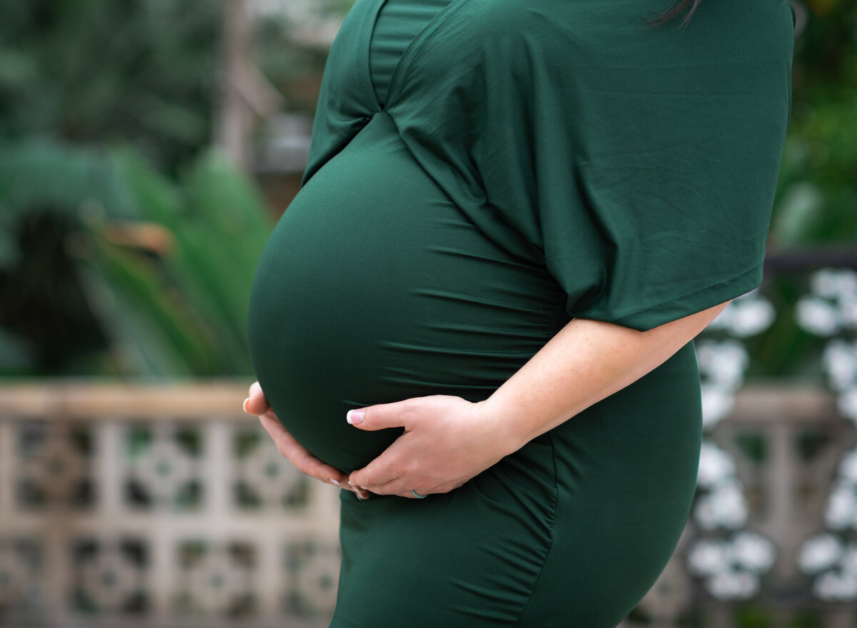 Close-up of a pregnant belly in a fitted green dress, hands gently cradling it