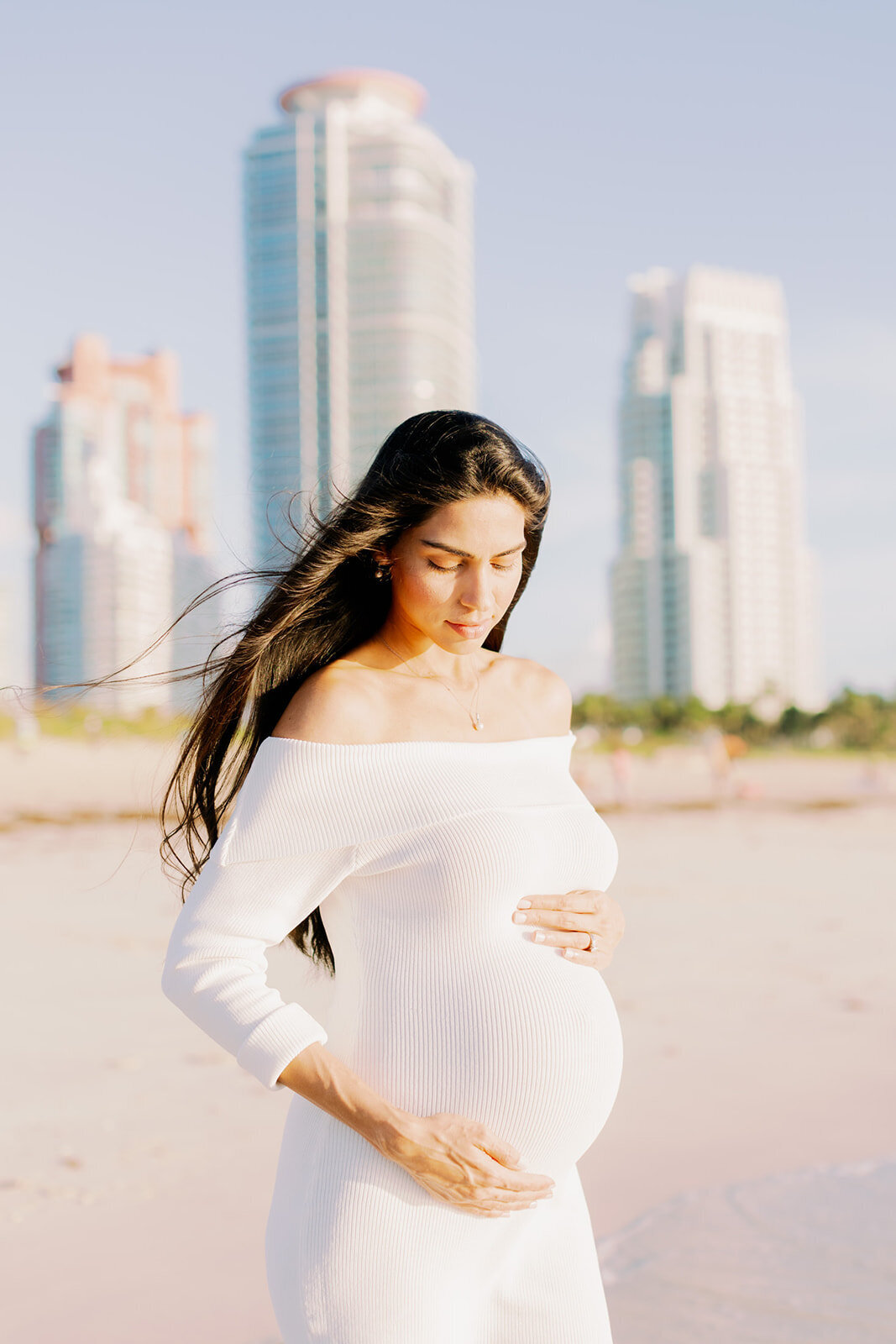 A pregnant woman with flowing hair stands near the ocean, skyscrapers in the background.
