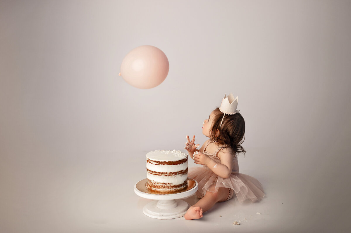 one year old baby girl with long  brown hair  wearing pink tutu and birthday hat sitting with a beautiful  neutral tiered cake with frosting while llooking up at a single pale pink balloon