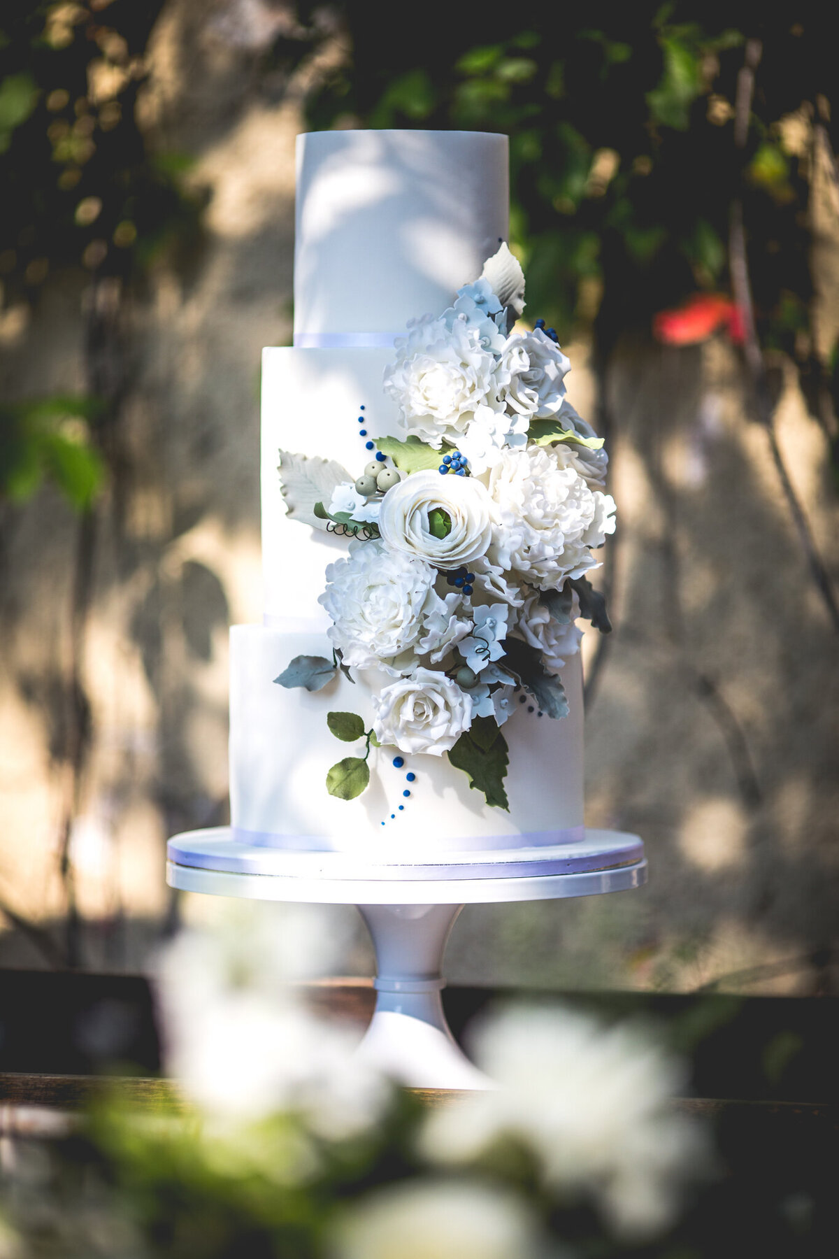 Elegant and romantic, three-tiered wedding cake with blue and purple  cascading florals, by Yvonne's Delightful Cakes, classic cakes & desserts in Calgary, Alberta, featured on the Brontë Bride Vendor Guide.