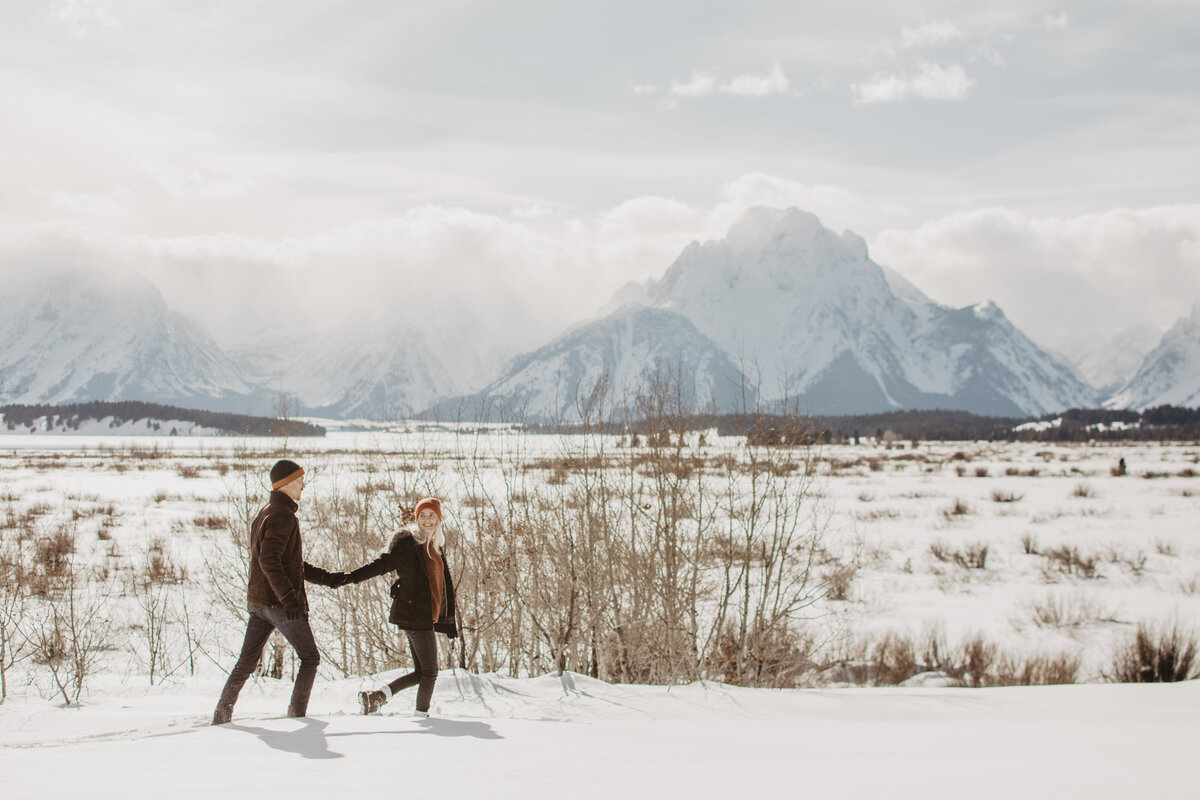 jackson hole photographers captures woman holding her fiances hands as she guides his through a snowy field in front of the Tetons in Jackson Hole for their winter engagement session