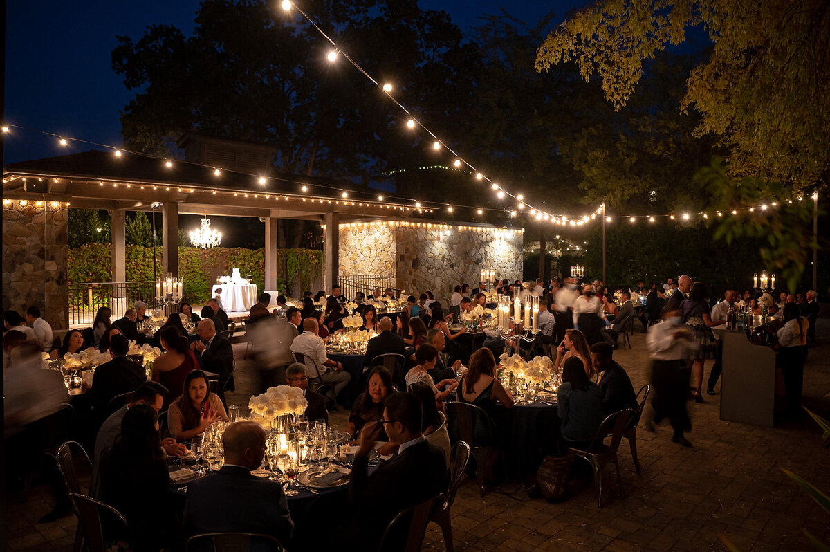 San Francisco Bay Area wedding photographer documents evening reception celebration with guests dining under romantic string lighting and stone architecture at Estate Yountville in Napa Valley