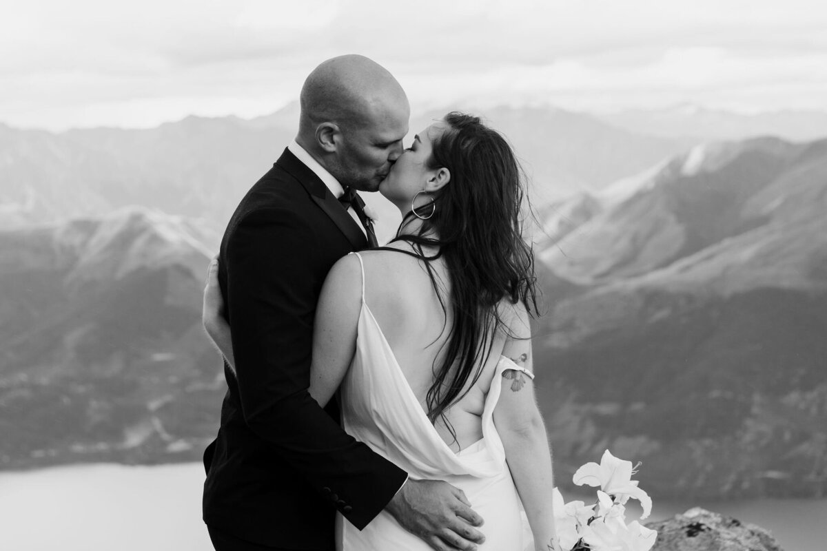 The Lovers Elopement Co - couple kiss on top of mountain with lake below, heli wedding