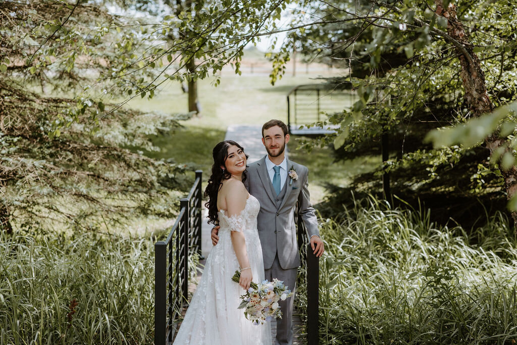 a bride in a flowing white gown and groom in a gray suit with blue tie on the bridge in the gardens of Willowbrook wedding venue