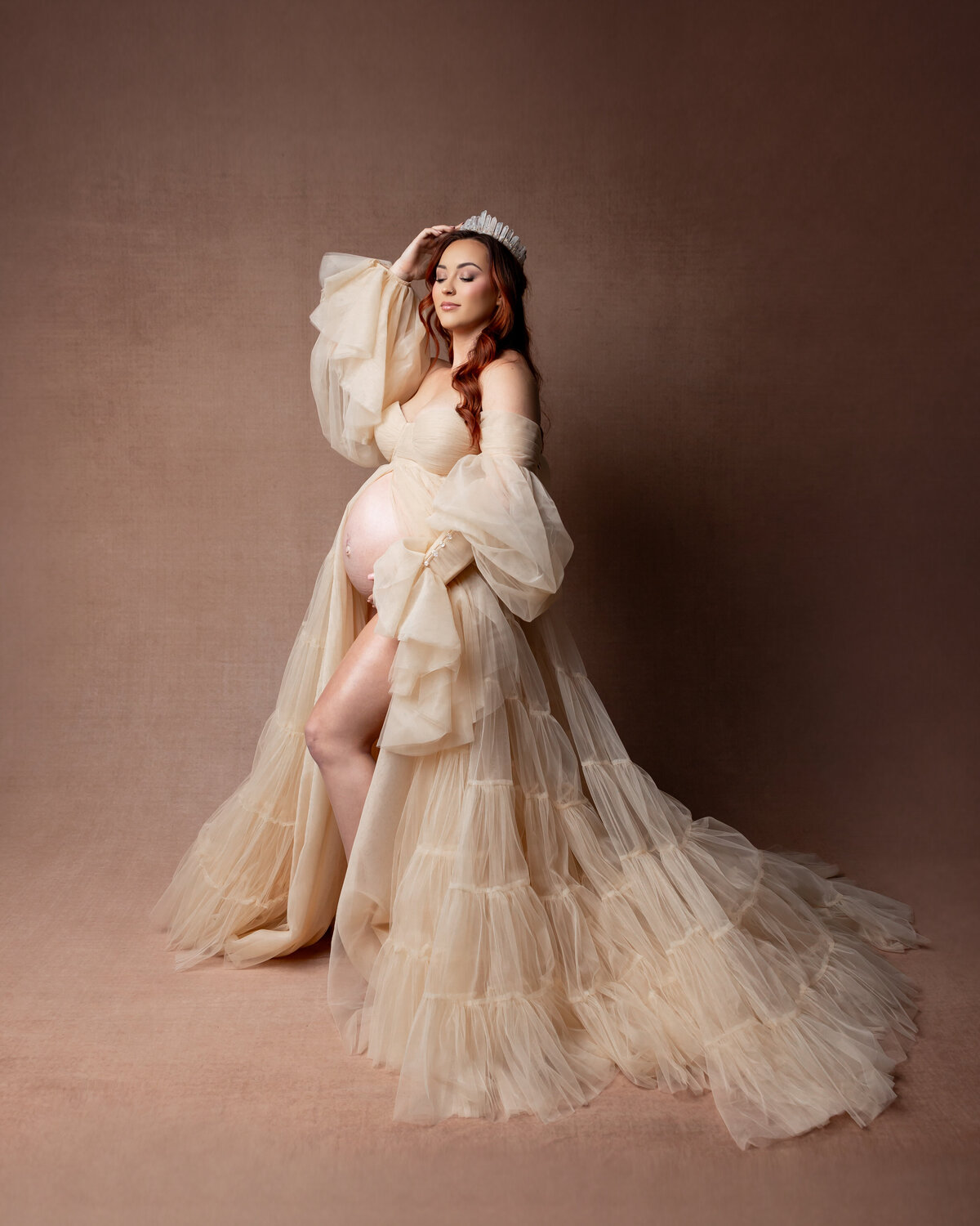 The expectant mother, wearing a dusty white dress and a delicate crown, poses gracefully against a warm brown backdrop. Captured by Inland Empire maternity photographer Kaitlyn Dawn Photography.