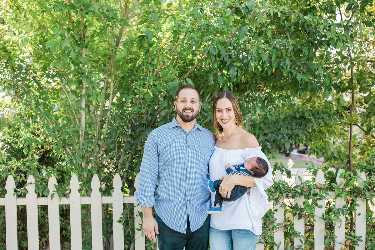 Young couple pose for photos while holding their infant child in front of a white picket fence and green tree