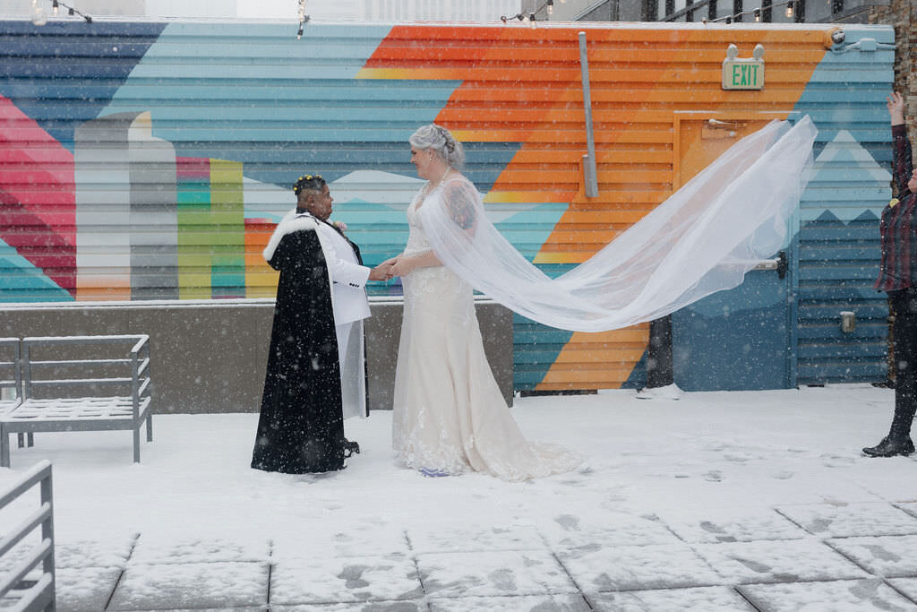 A wedding couple standing outside in the snow while one of their veils flows in the wind.