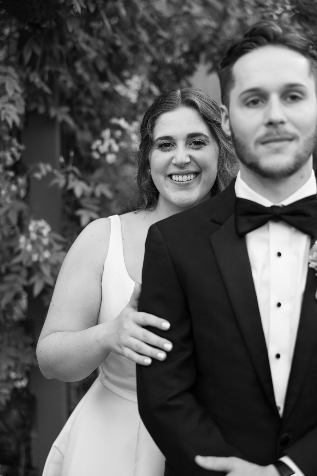 A bride standing behind a groom smiling with one hand on his arm