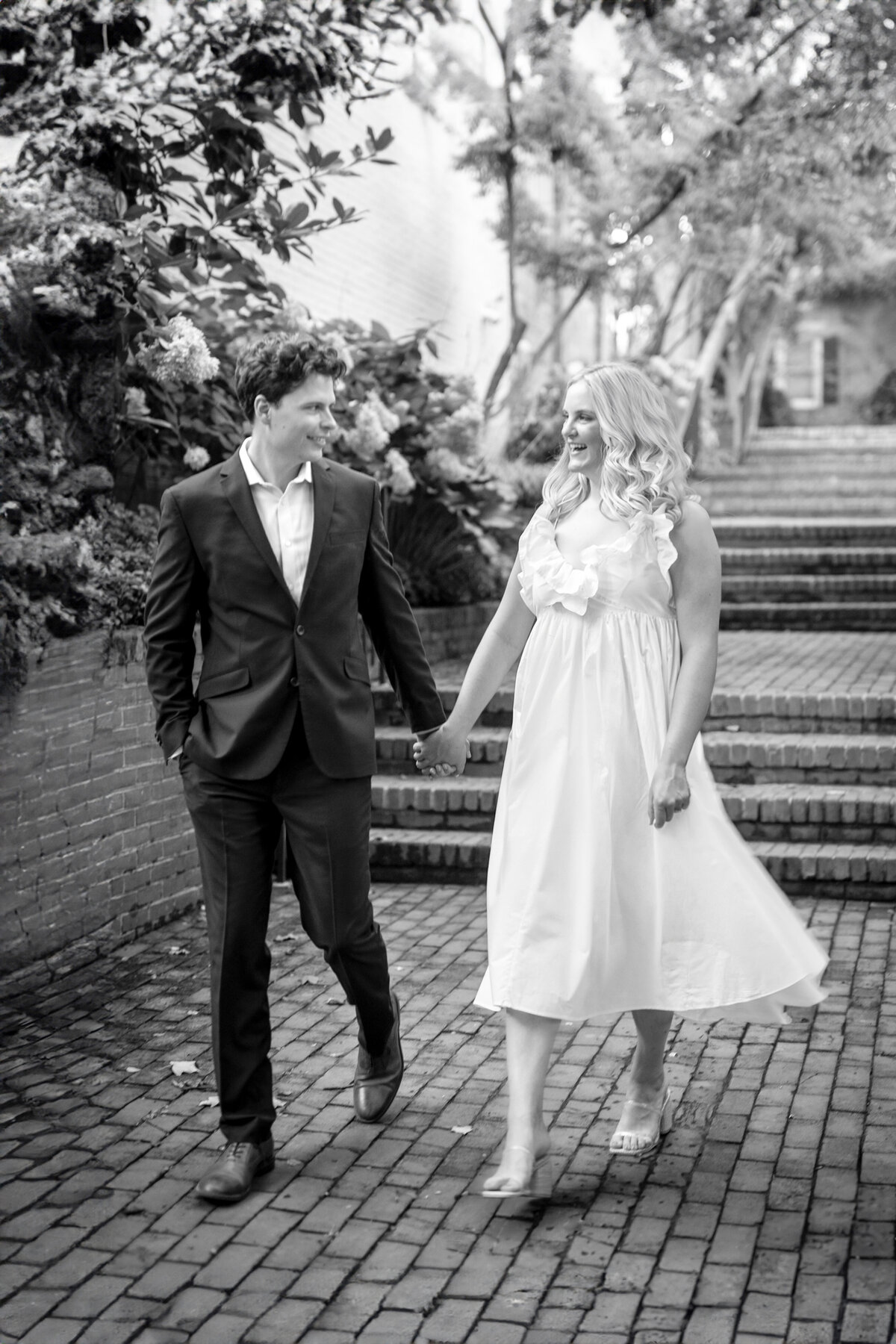 A couple walks hand in hand along a brick pathway. The man is wearing a suit, and the woman is in a white dress. They are smiling and surrounded by greenery and trees. The image is in black and white.