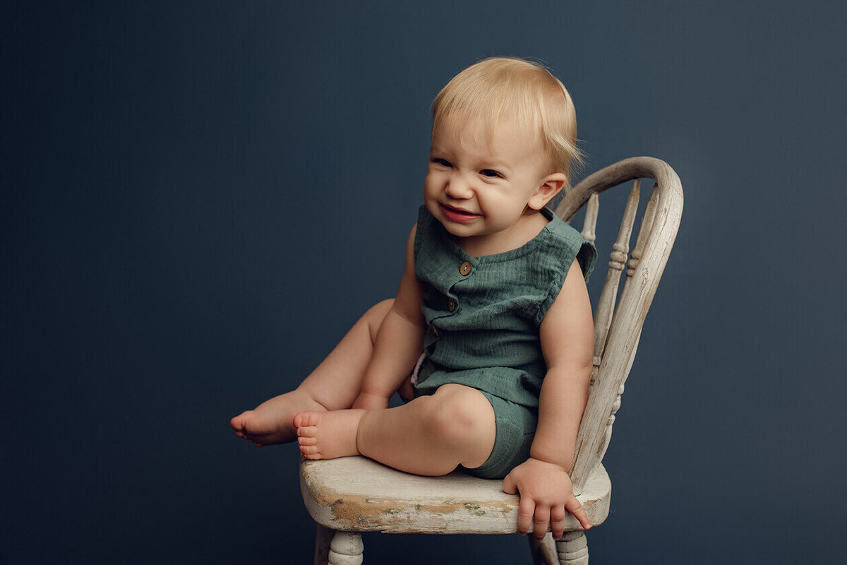 a one year old boy on a white chair with a blue backdrop smiling