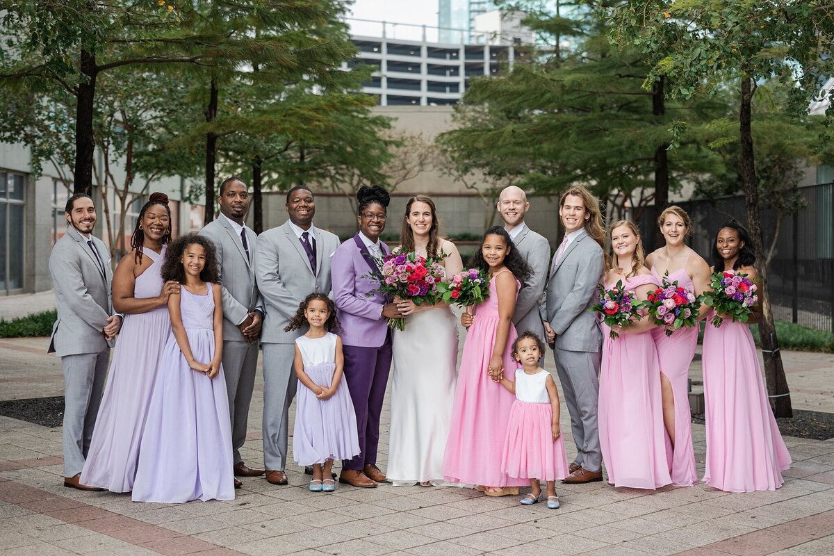 A formal portrait of two brides and their entire wedding party at the Westin Dallas Downtown in Dallas, Texas. The bride on the left is wearing a purple suit and is helping the other bride hold her bouquet. This bride's side of the wedding party is wearing either grey suits with purple ties or light purple dresses. The bride on the right is wearing a long, white dress and is also helping hold the large bouquet. Her side of the wedding party is wearing either grey suits with pink ties or light pink dresses.
