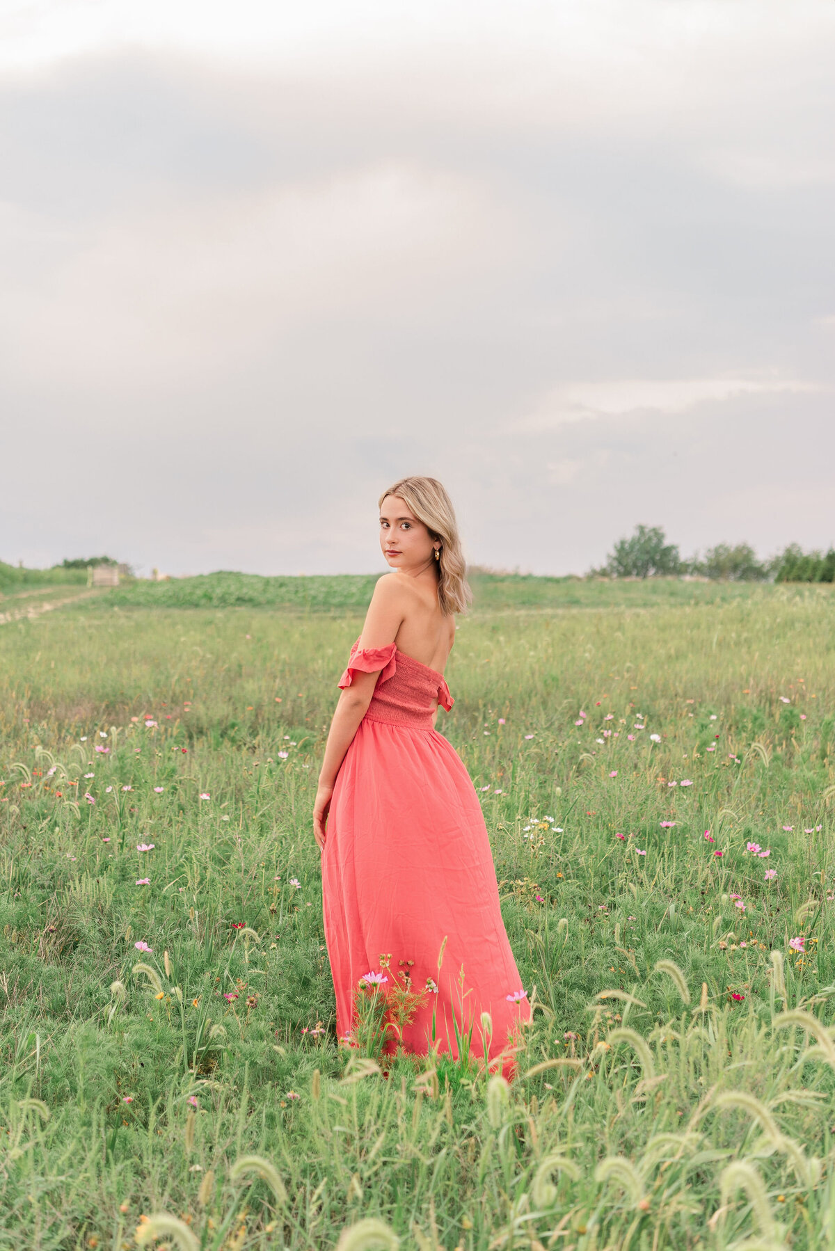 High school senior in coral dress standing in a wildflower field under a cloudy sky — Pittsburgh senior photographer Bree Stewart of Lens Love Photography.