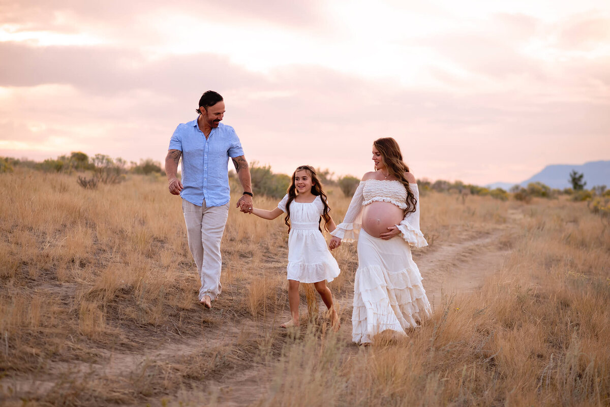 A family of three holds hands while walking towards the camera. They are smiling and there is a soft sunset in behind them and a grassy terrain. Taken in kamloops by Brittany Danielle
