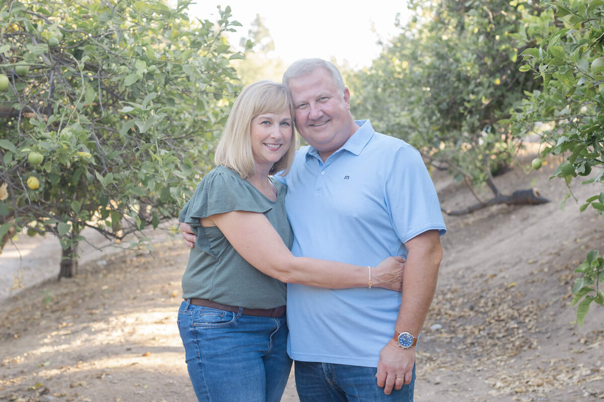A husband and wife with light blonde hair hugging each other's waists with their heads together smiling at the camera in a park with fruit trees.