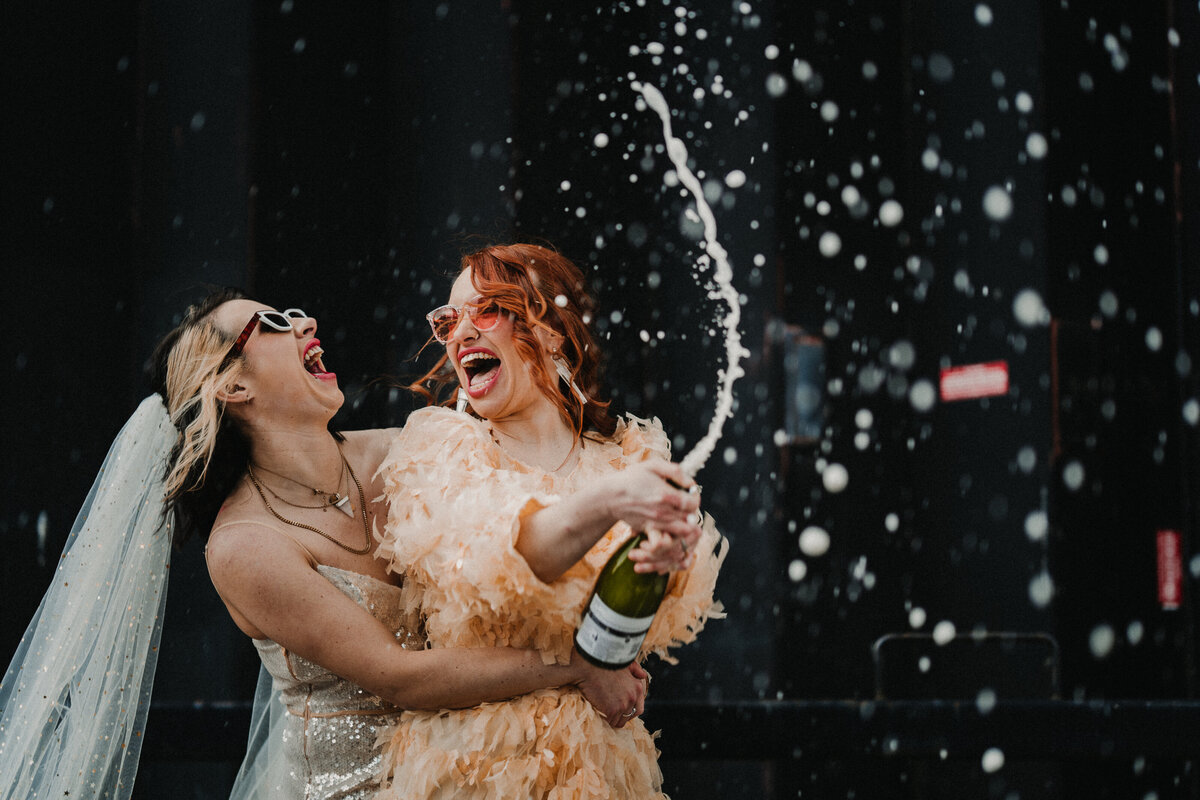 Fun wedding photography of two brides popping champagne