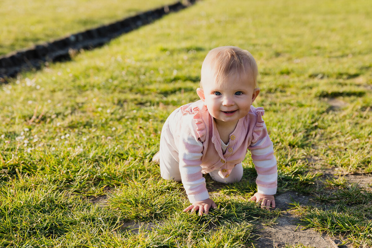 A baby girl crawling on grass, smiling at the camera, wearing a pink outfit. bright sunlight and shadow patterns on the ground.