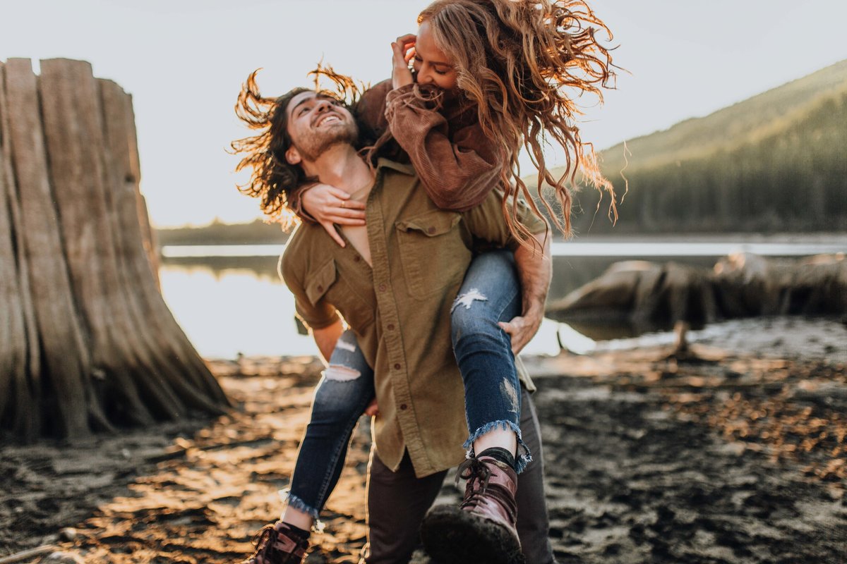 Married couple having fun during sunset session. Man giving woman a bouncy piggy back ride. Both have long hair and both hair is flying in different directions.