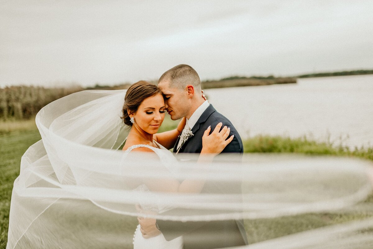 A bride and groom standing in front of a lake with her veil wrapped around them