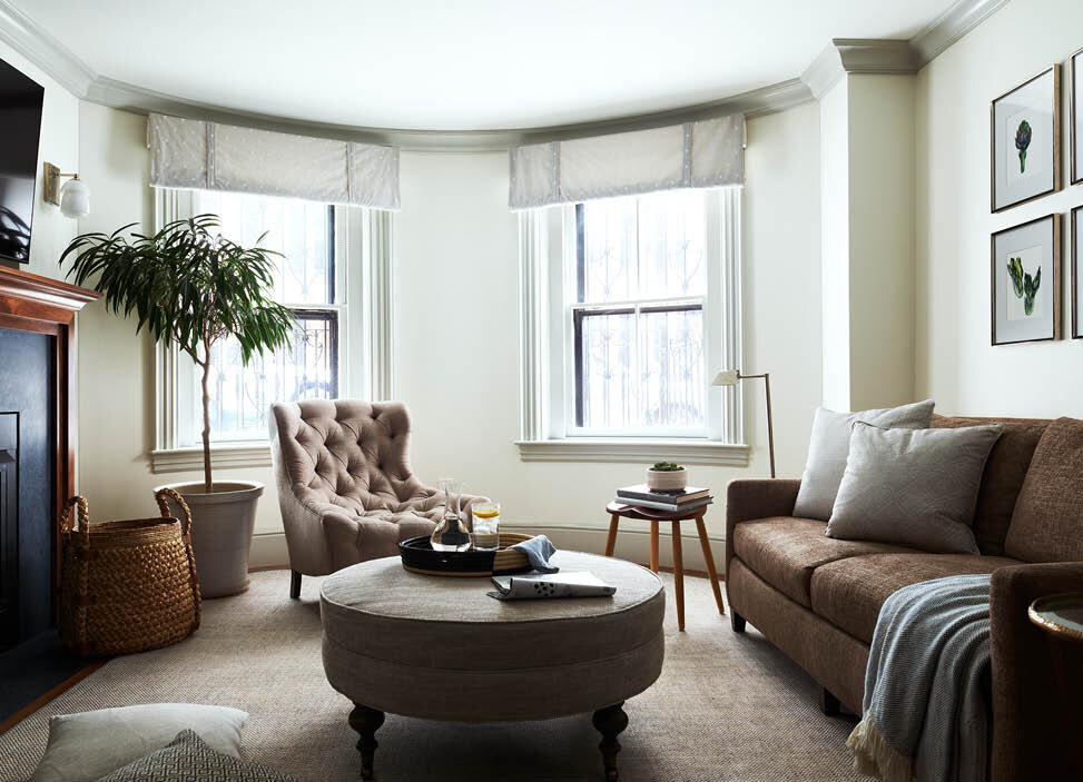 A simple fireplace faces a brown linen sofa, custom ottoman, and tufted lounge chair. Soft grey window treatments follow the brownstone's historic curves. By Sarah Scales.