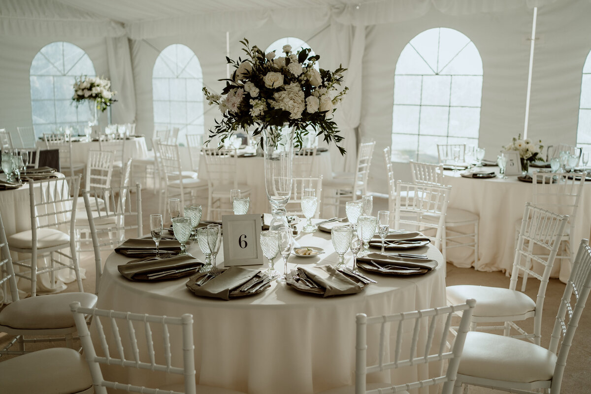 Elegant indoor wedding reception setup with round tables covered in white linens, decorated with tall floral centerpieces and place settings. White Chiavari chairs surround the tables, and the venue is filled with natural light from large windows.