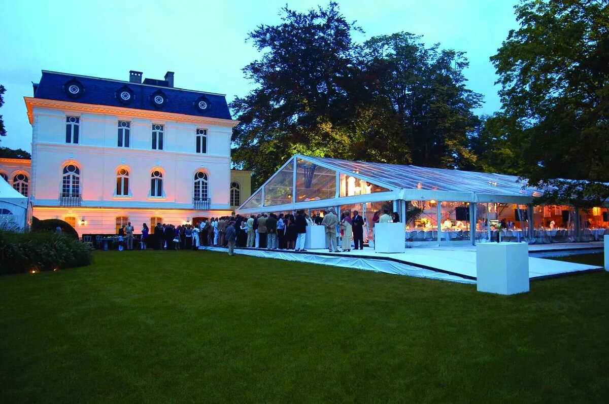 A large frame marquee with clear sides and roof in a large garden with a wedding party in full swing