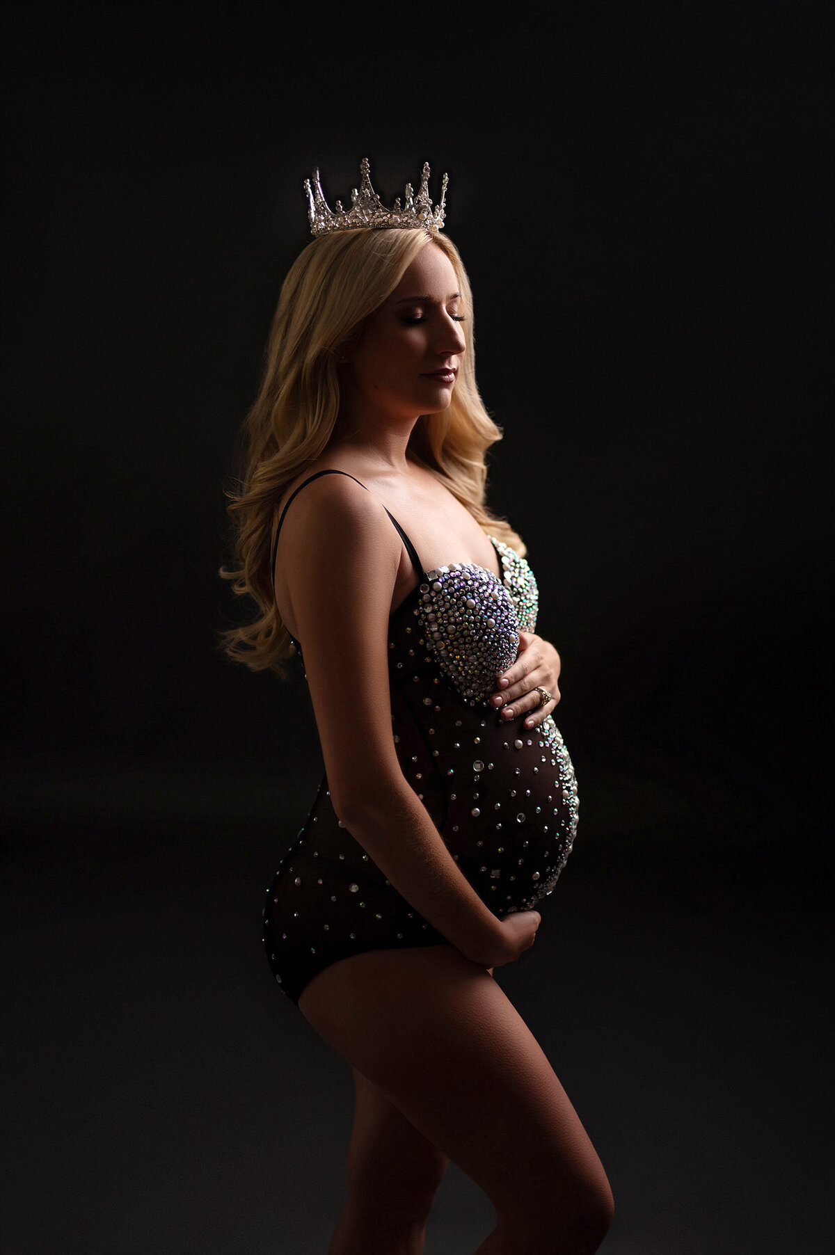 Pregnant woman standing in our Waukesha studio softly crading her belly while wearing a sheer, black maternity bodysuit adorned with pearls and crystals. A matching crown sits on top of her head as well.