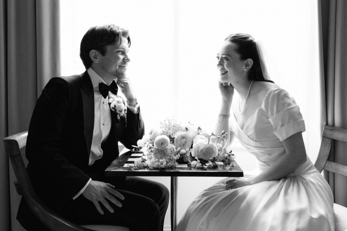 A timeless black and white portrait of a bride and groom sharing a quiet, intimate moment at a small table. Elegant florals and soft smiles capture the essence of a classic love story.