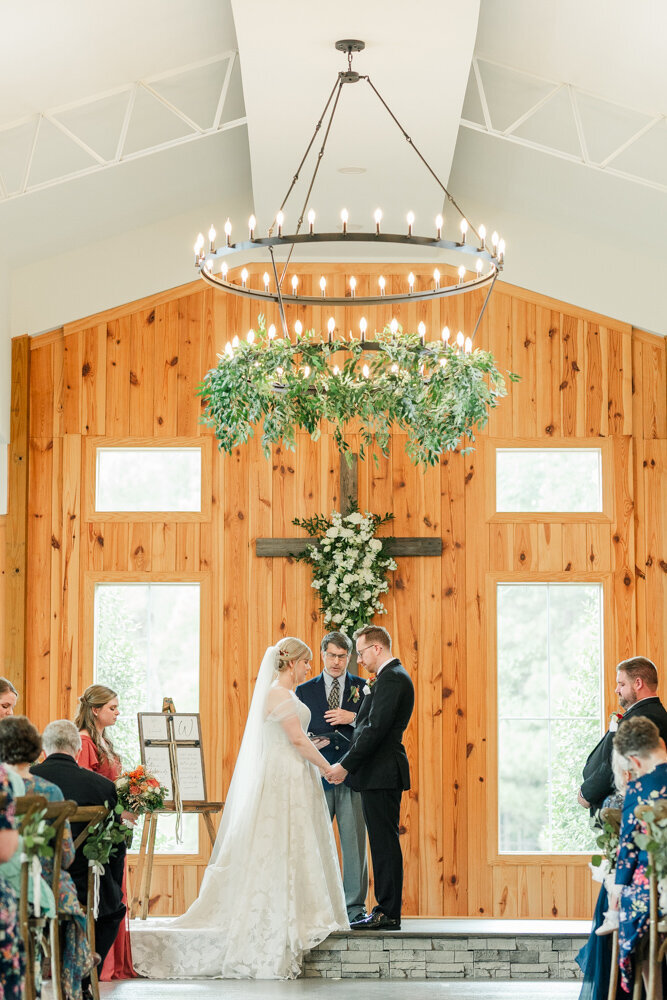 Bride and groom standing in front of a cross during their wedding ceremony, celebrating their sacred union surrounded by love and blessings.