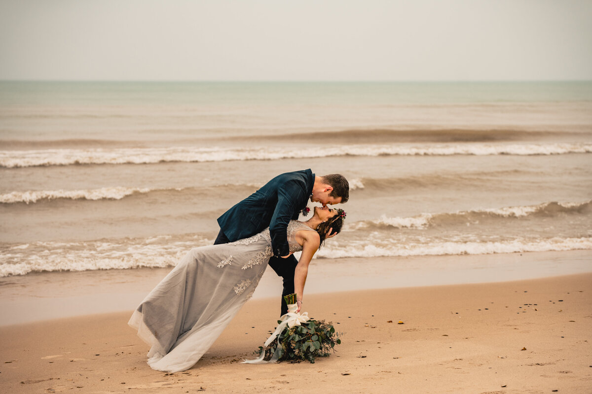 Wearing a silver wedding gown, the groom dips her in the pouring rain during this epic rainy beach elopement