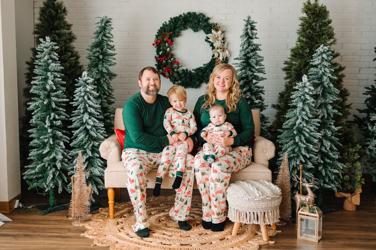 A family is taking a christmas holiday mini session photo . They are sitting on the coach and behind them are  christmas  trees with fake snow. They are dressed with green and white pajamas