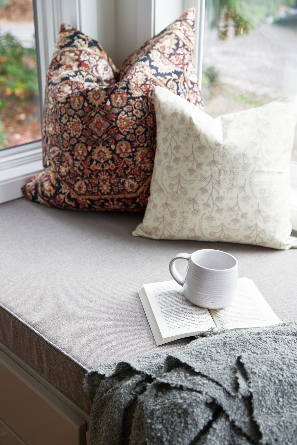 Window seat with brown wooden base with 4 drawers and silver knobs. Taupe cushion on top with a green and gray blanket, an open book and a cup of coffee. 3 throw pillows in the corners that are floral and striped.