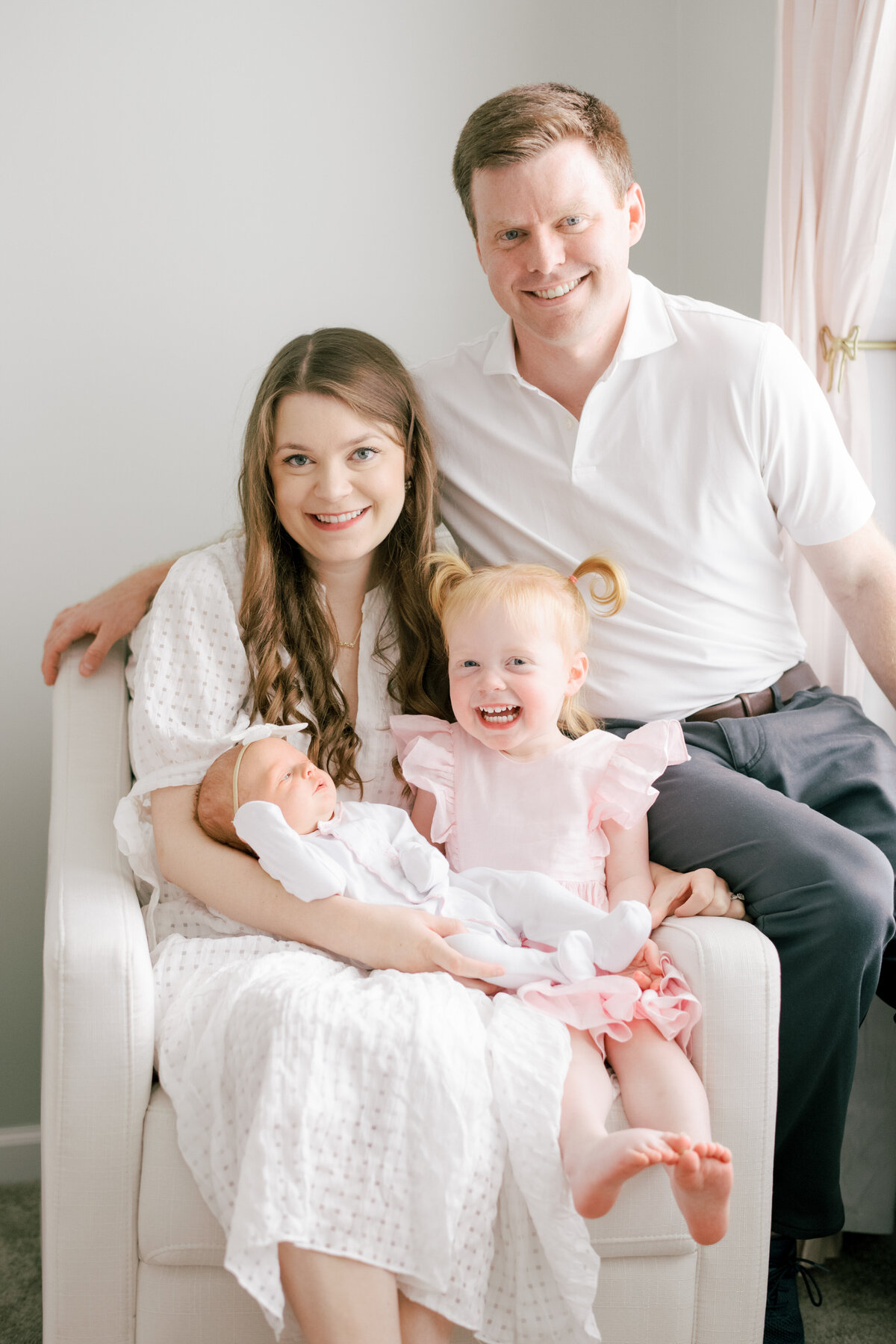 Big sister laughs as she sits on her parents lap for a photo with her new sister. Image by Raleigh newborn photographer A.J. Dunlap Photography.