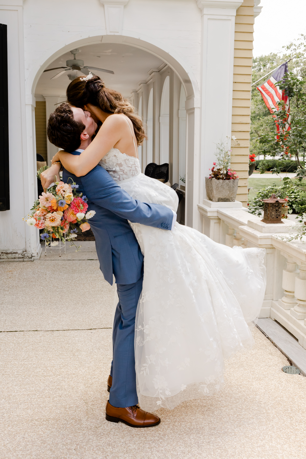 A groom joyfully lifts his bride for a kiss on the charming patio of the historic Lilac Inn in Brandon, Vermont, during their intimate wedding ceremony, surrounded by lush summer greenery and twinkling lights