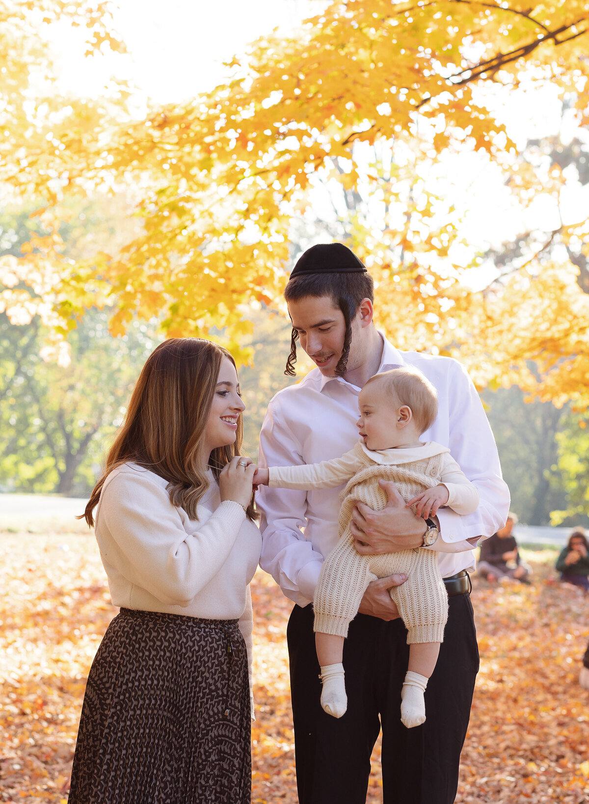 Mom dad and baby are standing in a brooklyn, ny park for a fall family photoshoot. Dad is holding baby and mom is holding baby's hand. Mom and dad are smiling at baby.  Captured by Brooklyn family photographer Chaya Bornstein